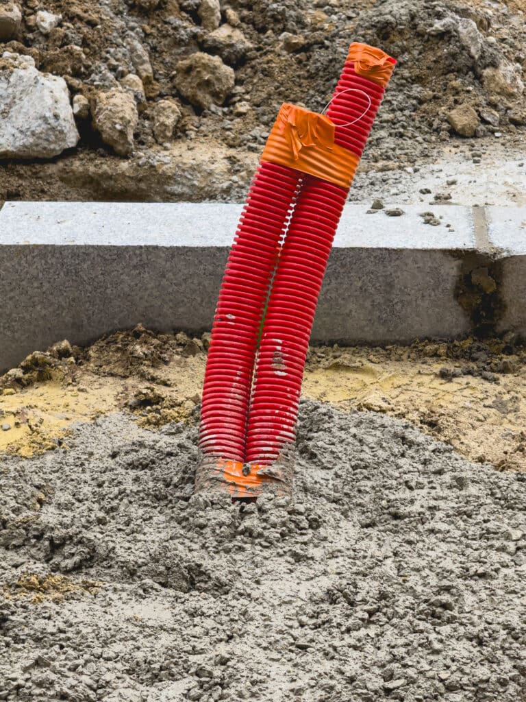Conduits rouges sur chantier: installation et travaux de bordure. Deux conduits rouges ondulés sortent de béton frais sur un chantier, avec une bordure en granit et de la terre en arrière-plan.