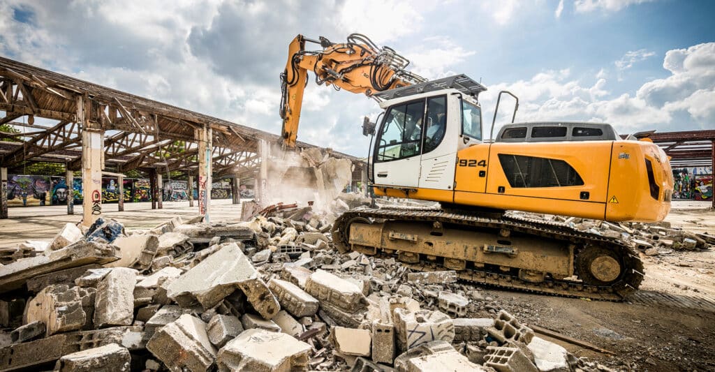 Excavatrice jaune Liebherr 924 démolissant une structure en bois sur un site jonché de gravats, avec des murs graffés et un ciel nuageux.