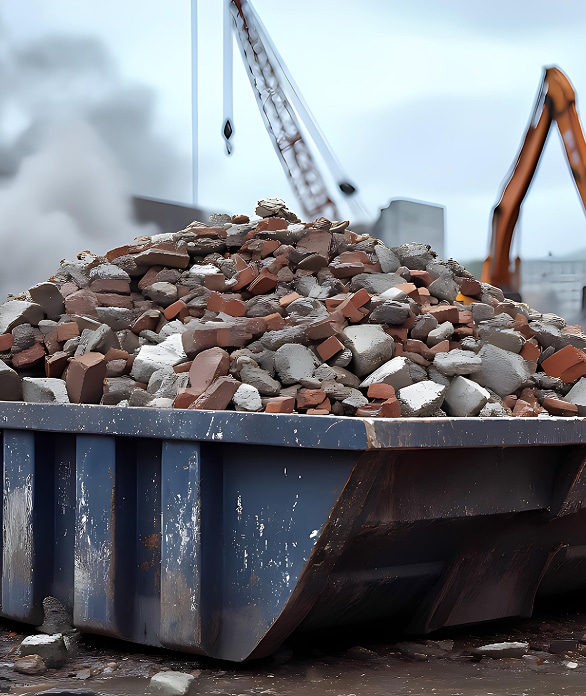 Gravats de démolition: benne, briques, béton sur chantier Benne à gravats bleue remplie de briques et béton sur chantier de démolition. Grue et excavatrice en arrière-plan, fumée.