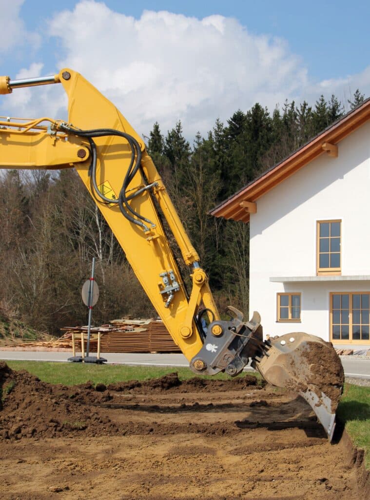 Pelle mécanique : Travaux de Terrassement pour Nouvelle Maison Grand bras de pelle mécanique jaune creusant la terre devant une maison neuve et des arbres. Un panneau de danger est visible sur l'engin.