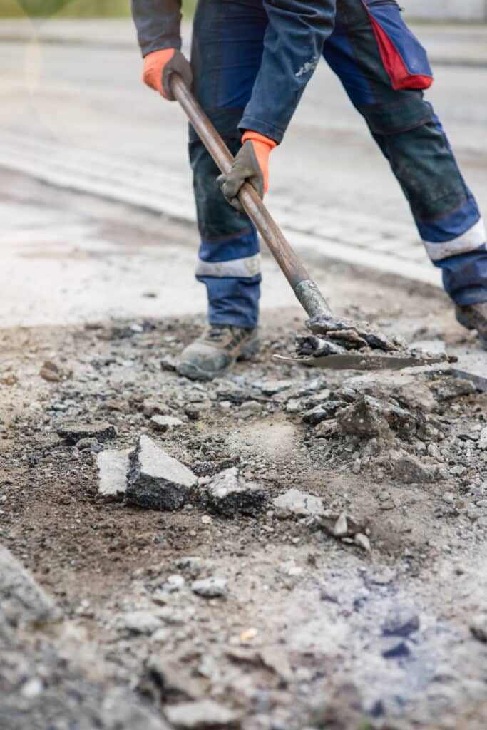 Réparation route : ouvrier pelle l'asphalte cassé Ouvrier en tenue bleue et gants orange pelle de l'asphalte cassé pour une réparation de route. Travail manuel de voirie.